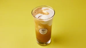 Freddo Cappuccino σε γυάλινο ποτήρι με πλούσιο αφρόγαλα, σε φόντο κίτρινο.